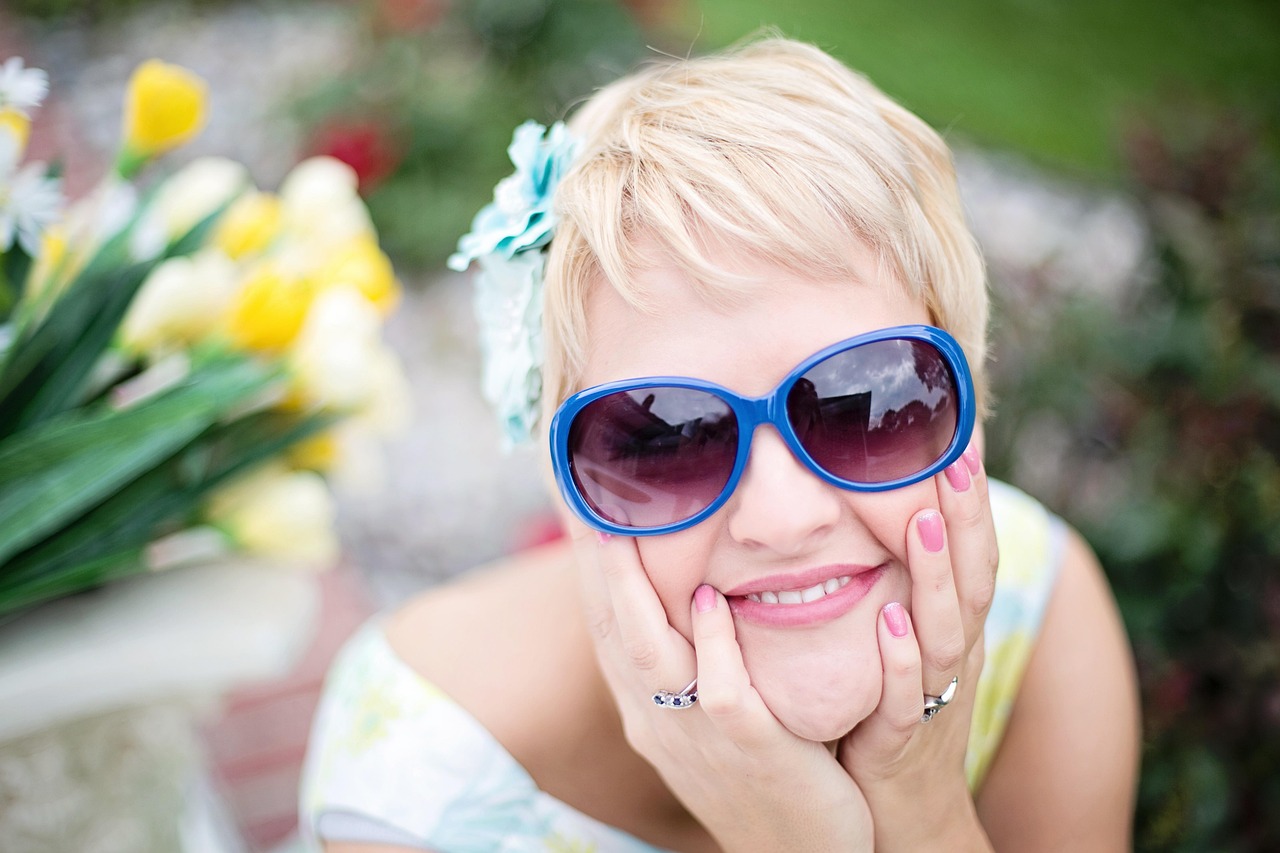A smiling woman in blue sunglasses, pink nails at her cheeks